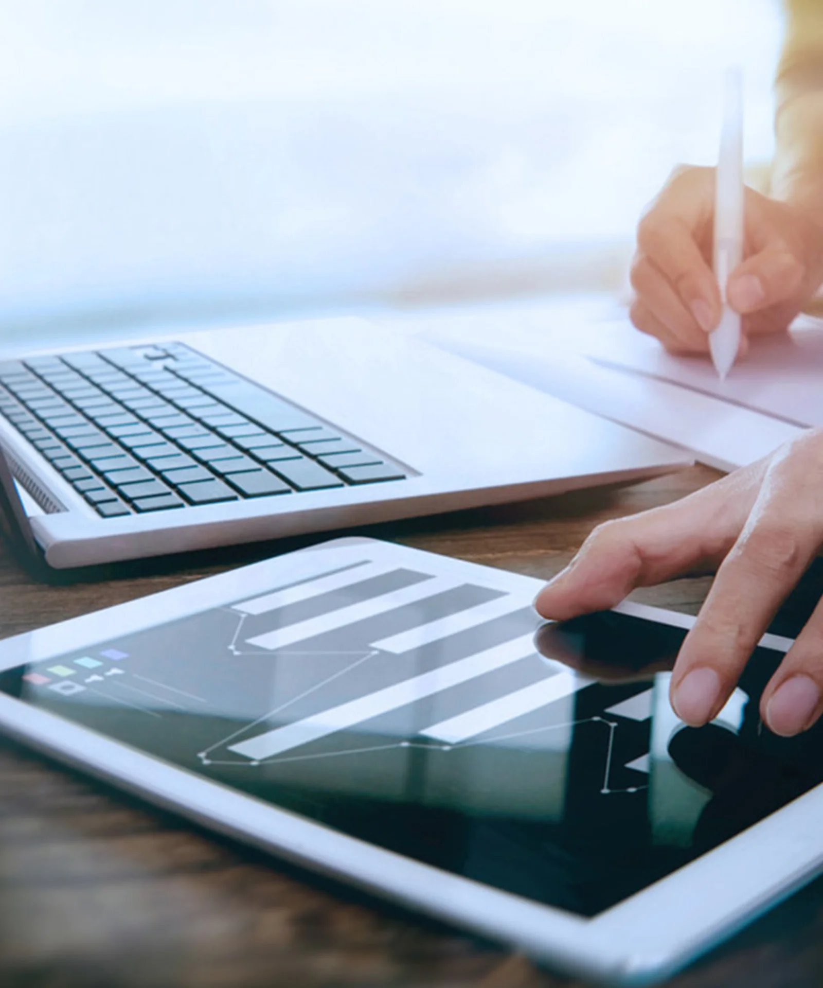A close-up of hands using a tablet to analyse data, with a laptop and a notepad in the background, symbolising the integration of digital tools in data-driven decision making.