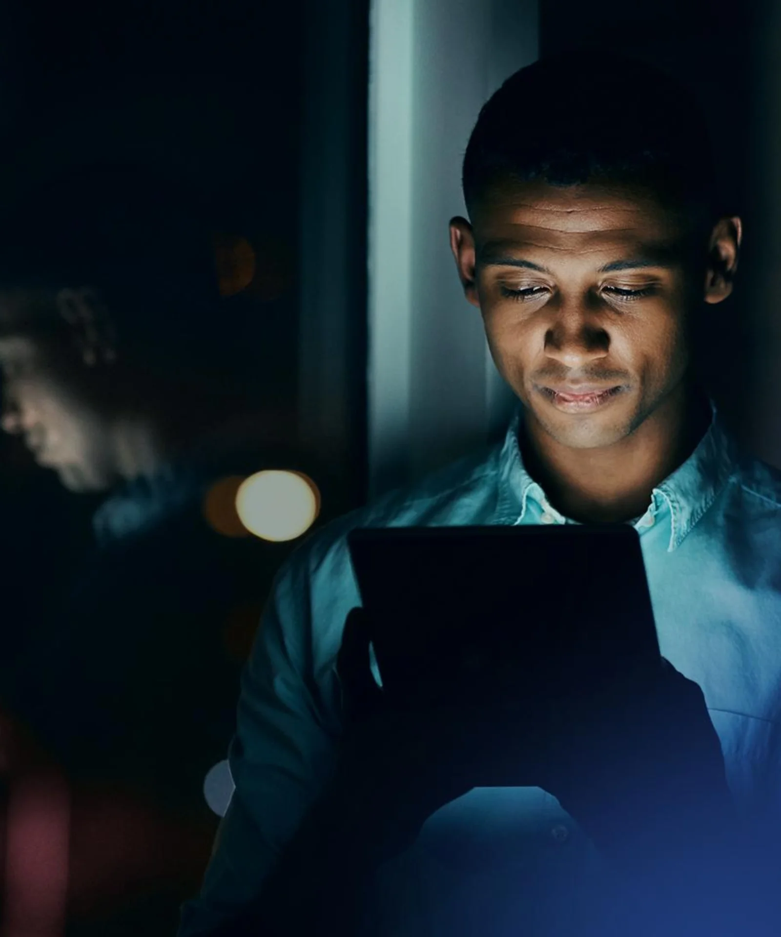 Man Using Tablet at Night by the Window Man looking at a tablet screen at night, standing by a window with city lights reflecting on the glass.