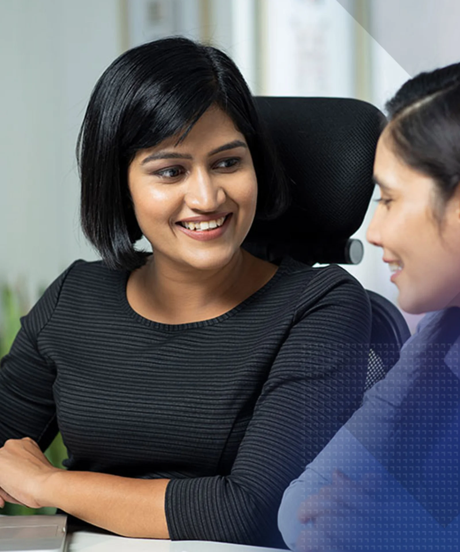 GFT Stage image - Culture and Values - India Two professional women engaged in a discussion at a desk, with a laptop open and one holding a pen, in a modern office environment.