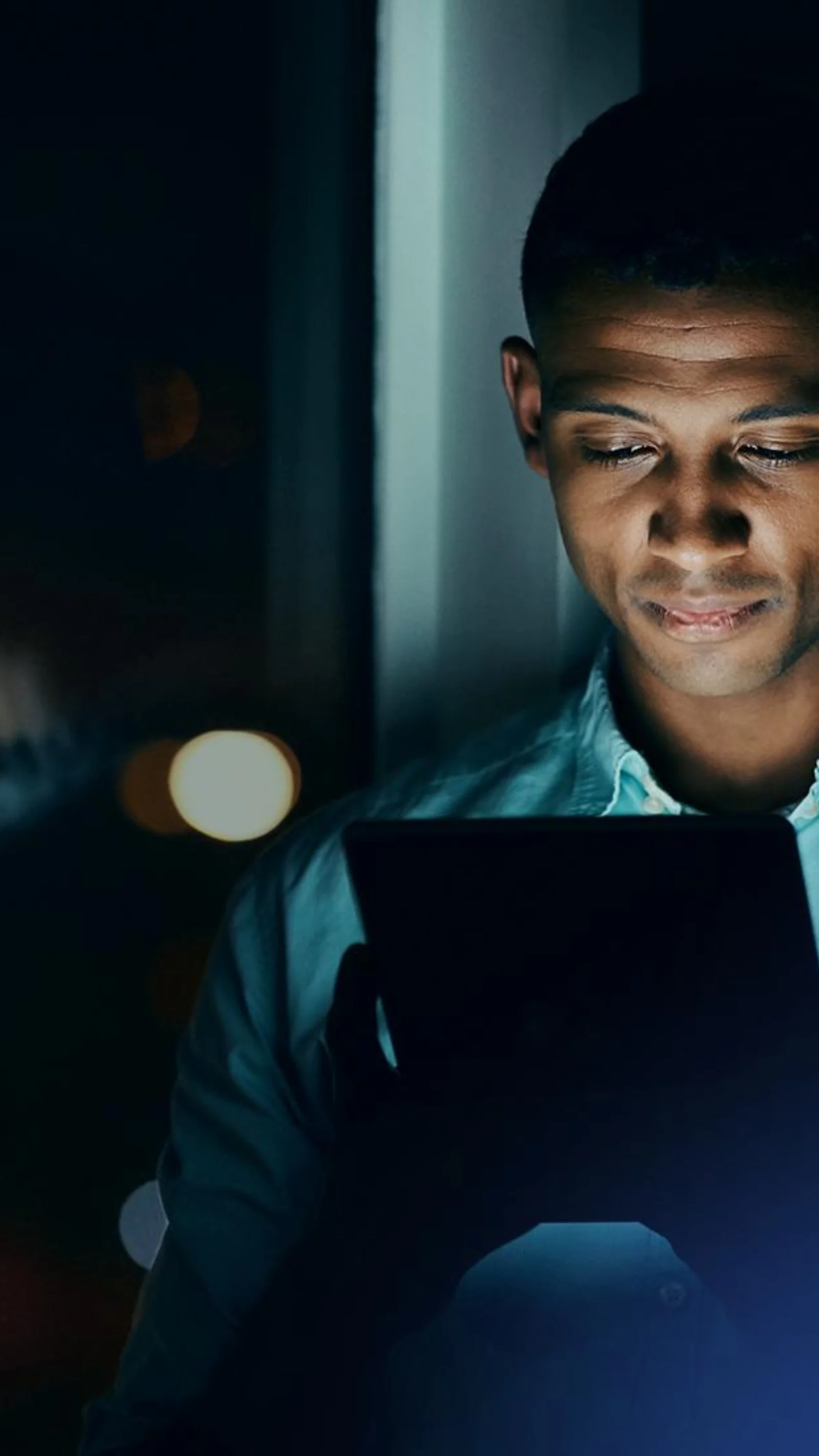 Man looking at a tablet screen at night, standing by a window with city lights reflecting on the glass.