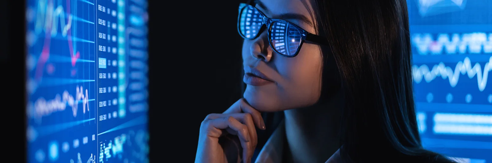 A woman in glasses, reflecting data charts, looks intently at multiple digital screens displaying various analytics and graphs.