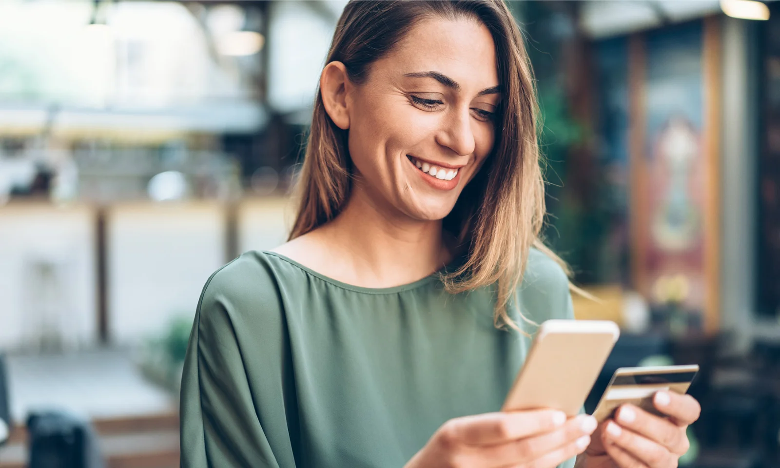 Comparing Open Banking APIs and Financial Aggregator APIs - 04 Smiling woman using a smartphone and holding a credit card for an online payment in a cafe.