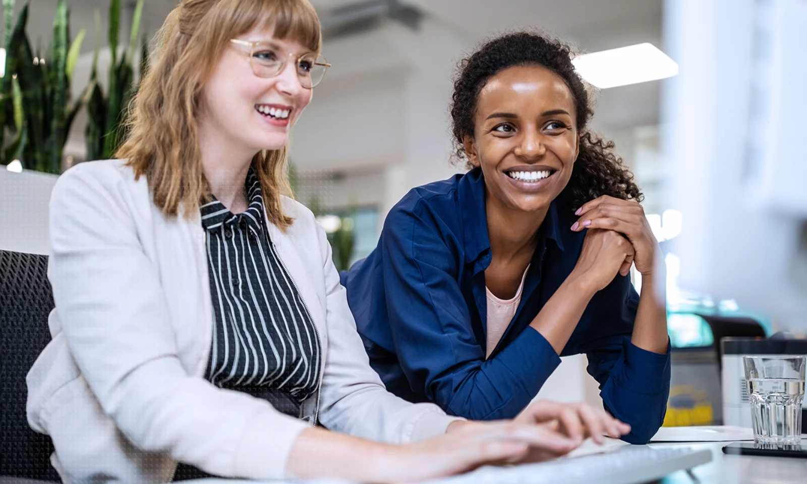 GFT Blog - AI as Thougth Partner - 01 Two professional women smiling and working together on a desktop computer in a modern office environment.