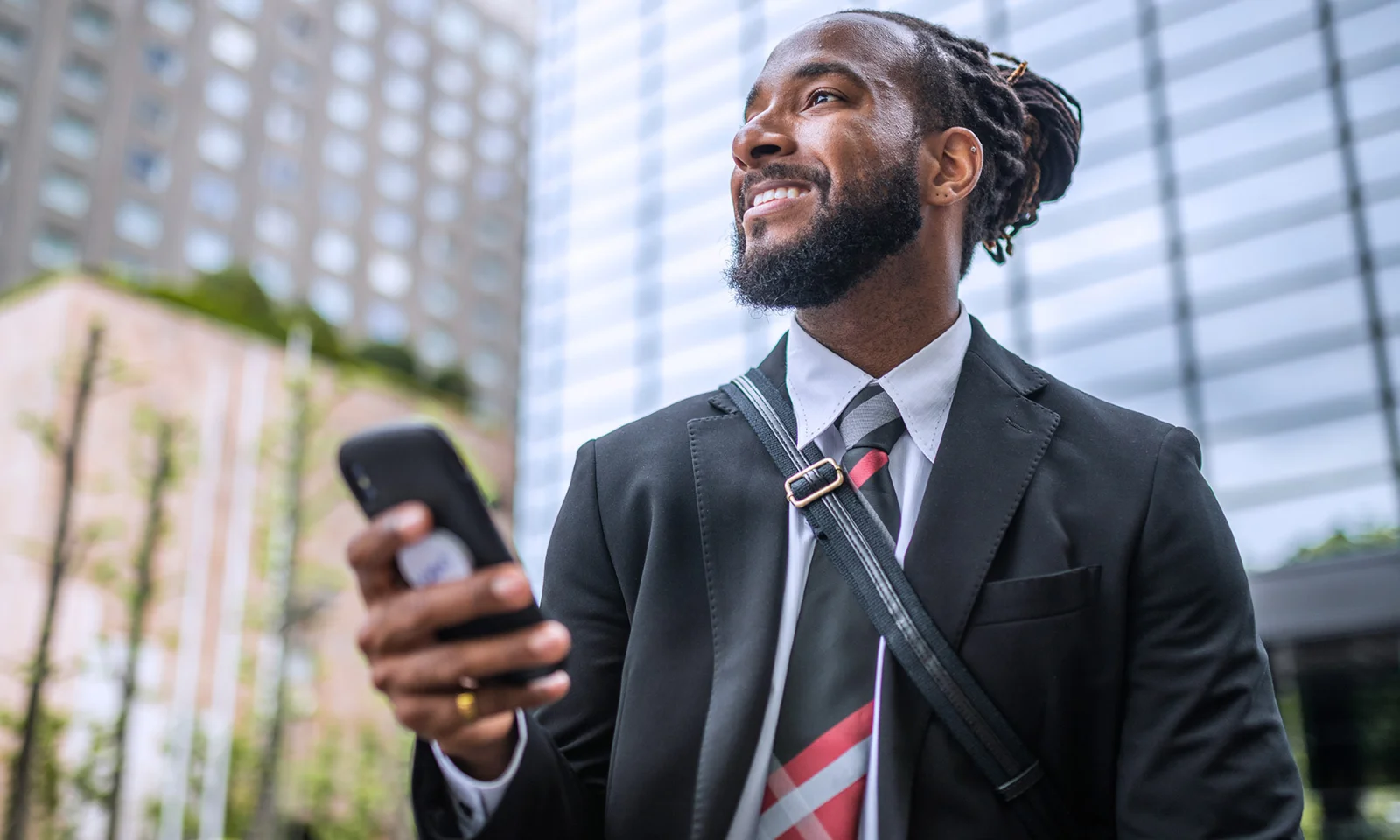 Comparing Open Banking APIs and Financial Aggregator APIs - 05 Smiling businessman in a suit holding a smartphone in a city environment with modern buildings in the background.