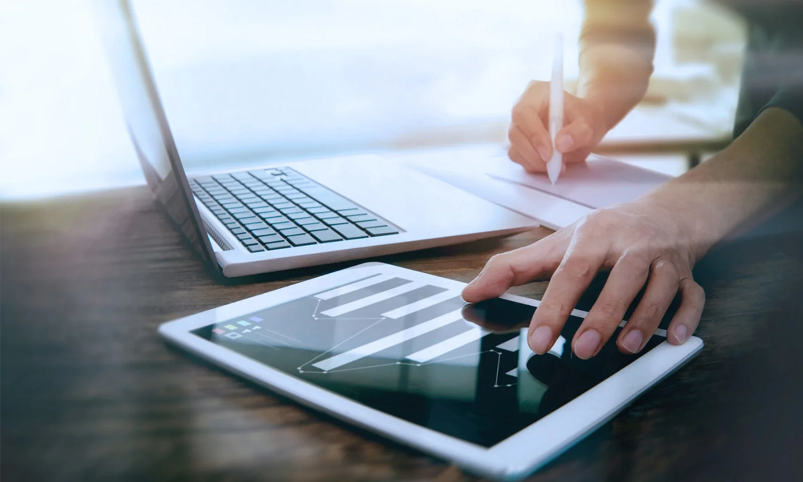 A close-up of hands using a tablet to analyse data, with a laptop and a notepad in the background, symbolising the integration of digital tools in data-driven decision making.