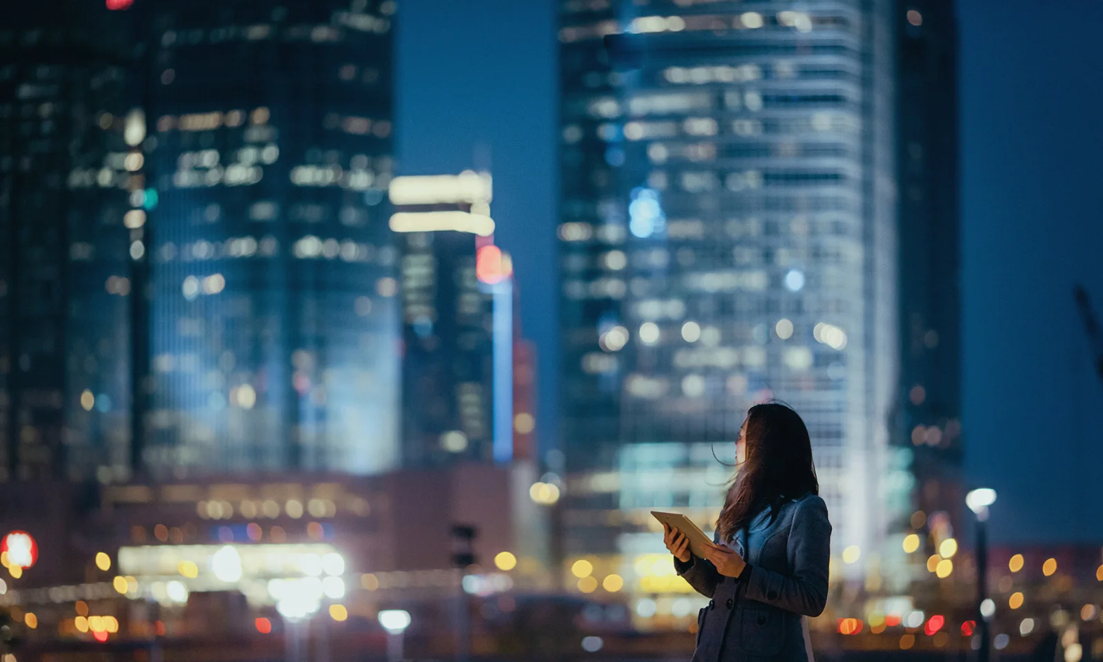 Una mujer que sostiene una tableta mira hacia los rascacielos iluminados de una ciudad por la noche, que simbolizan la innovación digital y la tecnología en un entorno urbano. Mercados de capitales visuales clave