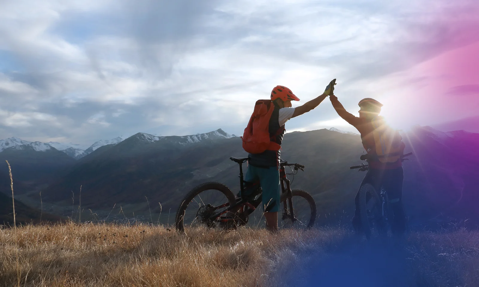 Zwei Radfahrer geben sich auf einem Berggipfel ein High-Five, im Hintergrund eine atemberaubende Landschaft aus Bergen und Tälern, was Teamwork und gemeinsame Leistung symbolisiert.