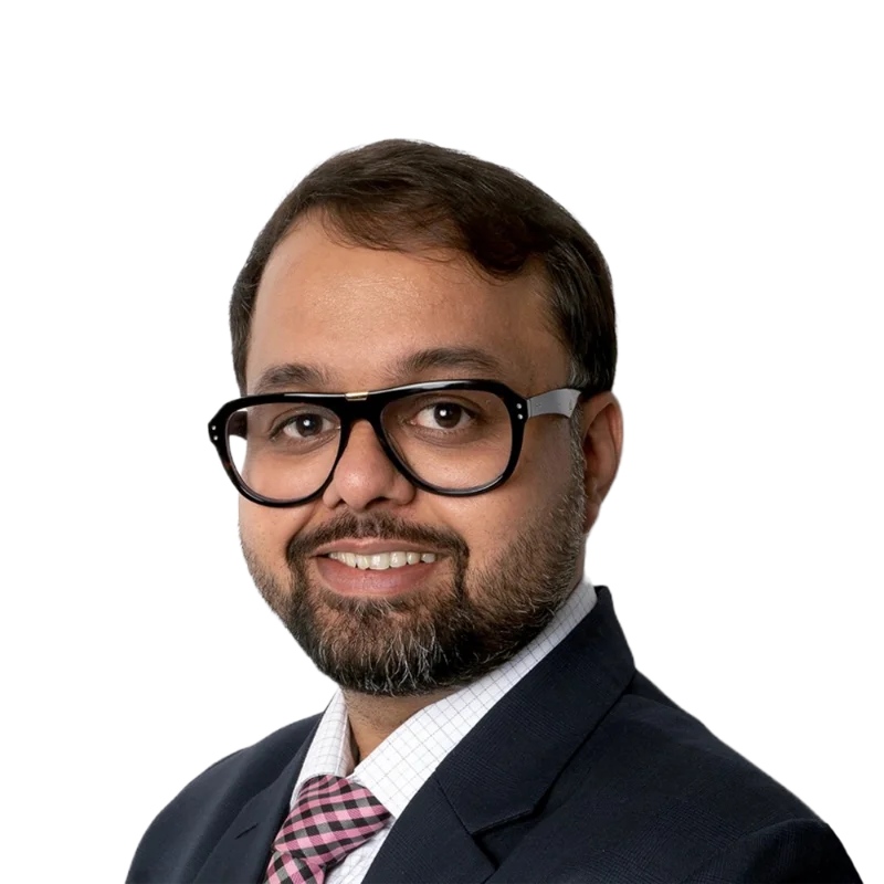 Portrait of Aaditya wearing glasses, a dark suit, white shirt, and a pink checked tie, smiling confidently.